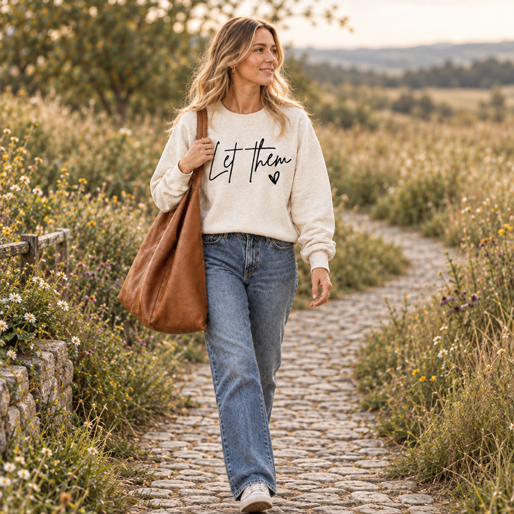 Woman in the Let Them sweatshirt walking along a cobblestone meadow path with a large tote